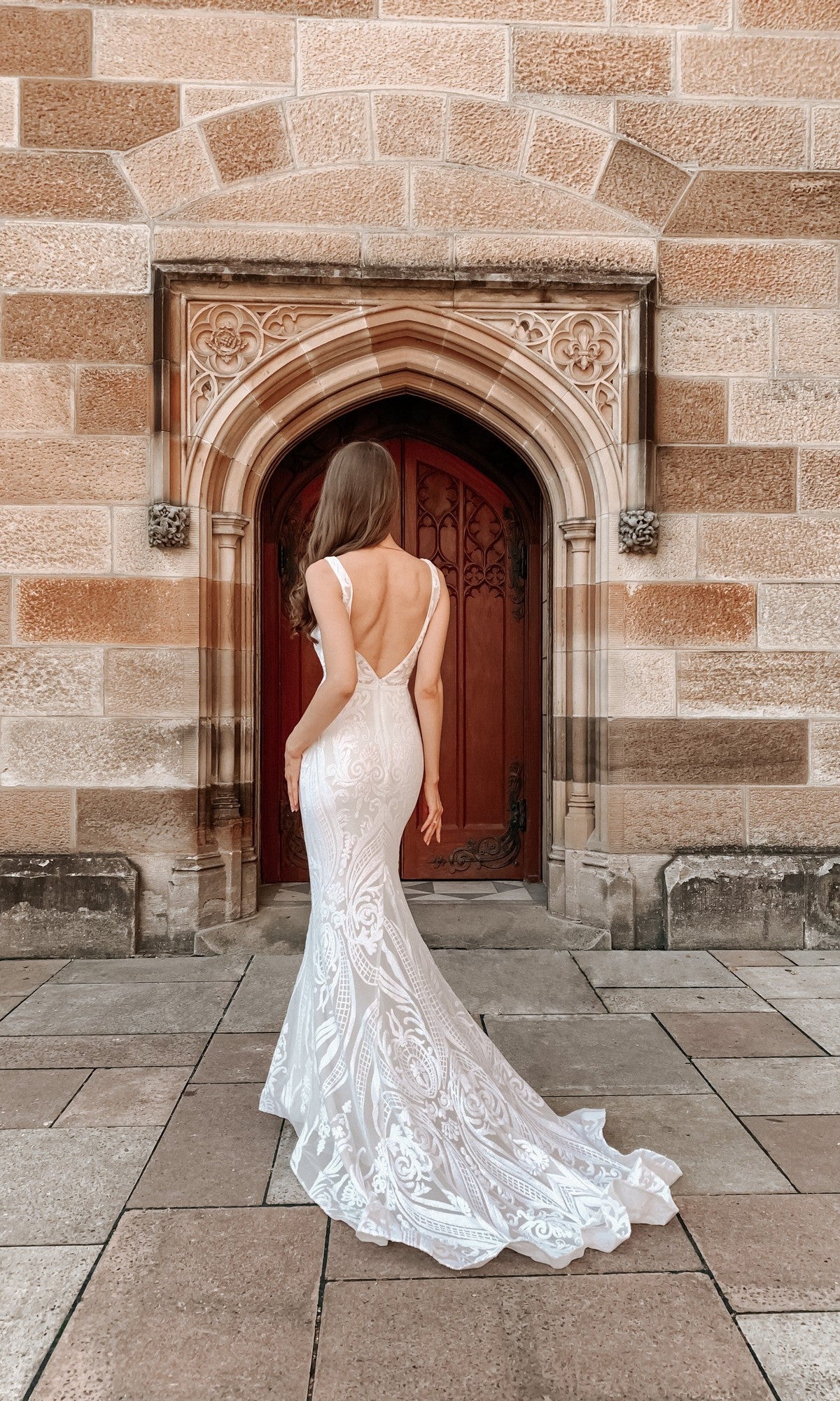 A woman in the White Lace Mermaid Wedding Gown: Tina Holly BA109 stands facing a wooden arched door set in a stone building.
